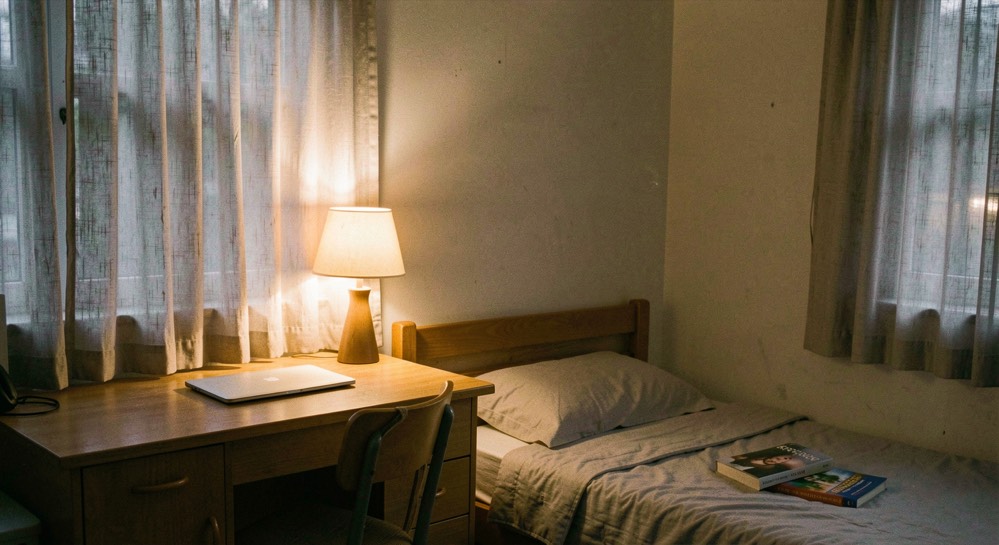 A small student bedroom in the evening with the curtains drawn, one warm lamp lit in the corner, a closed laptop on the desk and a book face down on the bed, no overhead light.