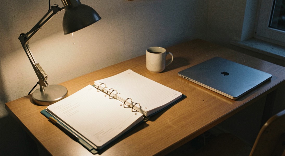 A student desk at night lit by one warm lamp, an open ring binder, a half full mug and a laptop closed to the side, wide shot taken slightly from above.