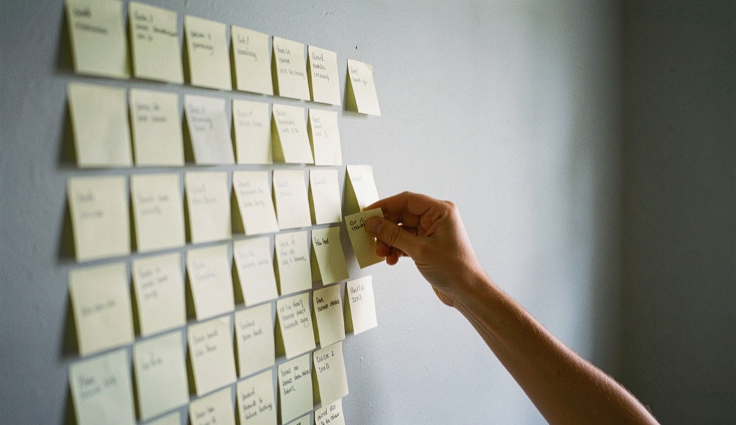 A wall covered with small square sticky notes arranged in columns, one hand moving a single note from one column to another, plain painted wall, cool afternoon light.